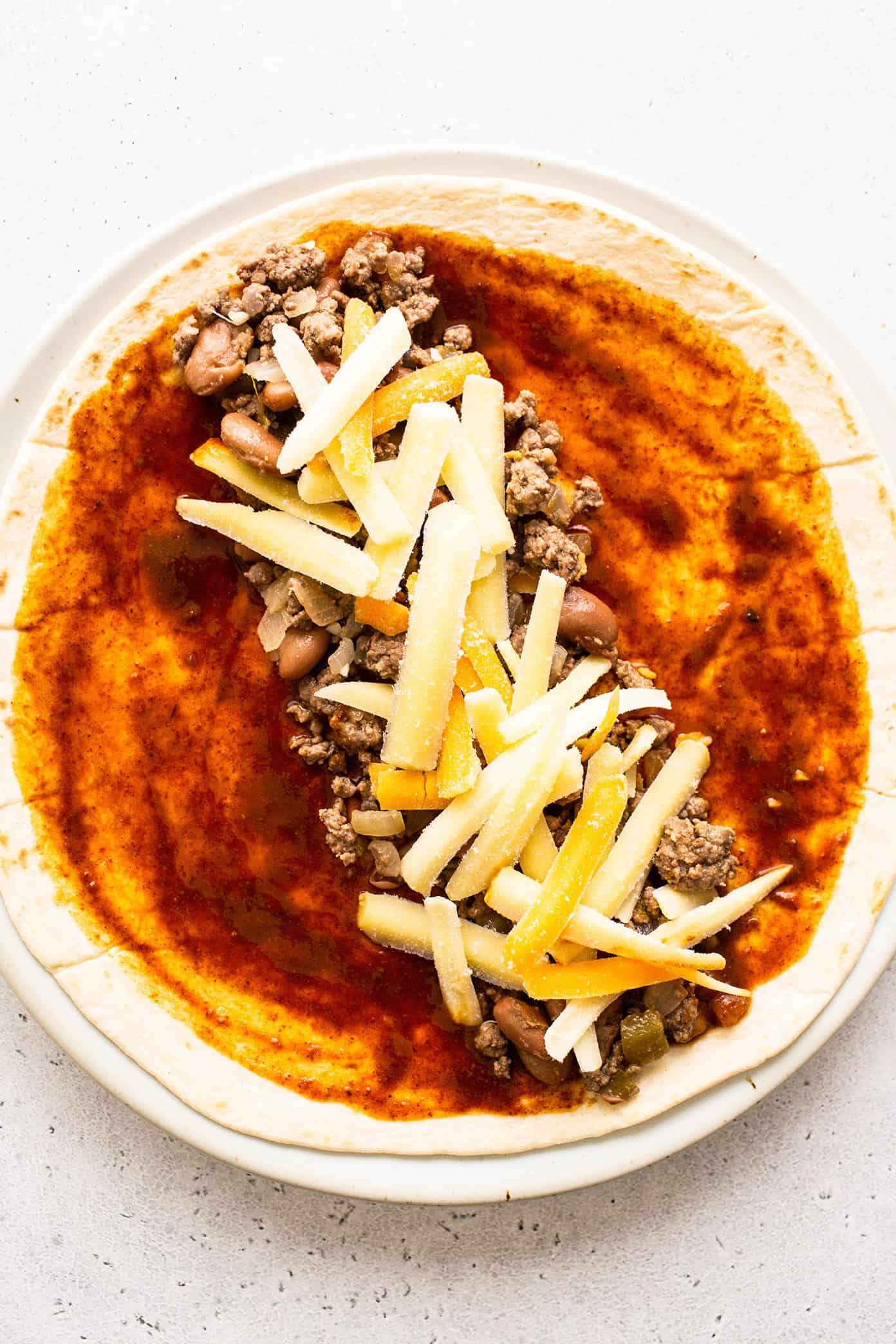 Beef enchilada filling spread on a tortilla with cheese and red enchilada sauce