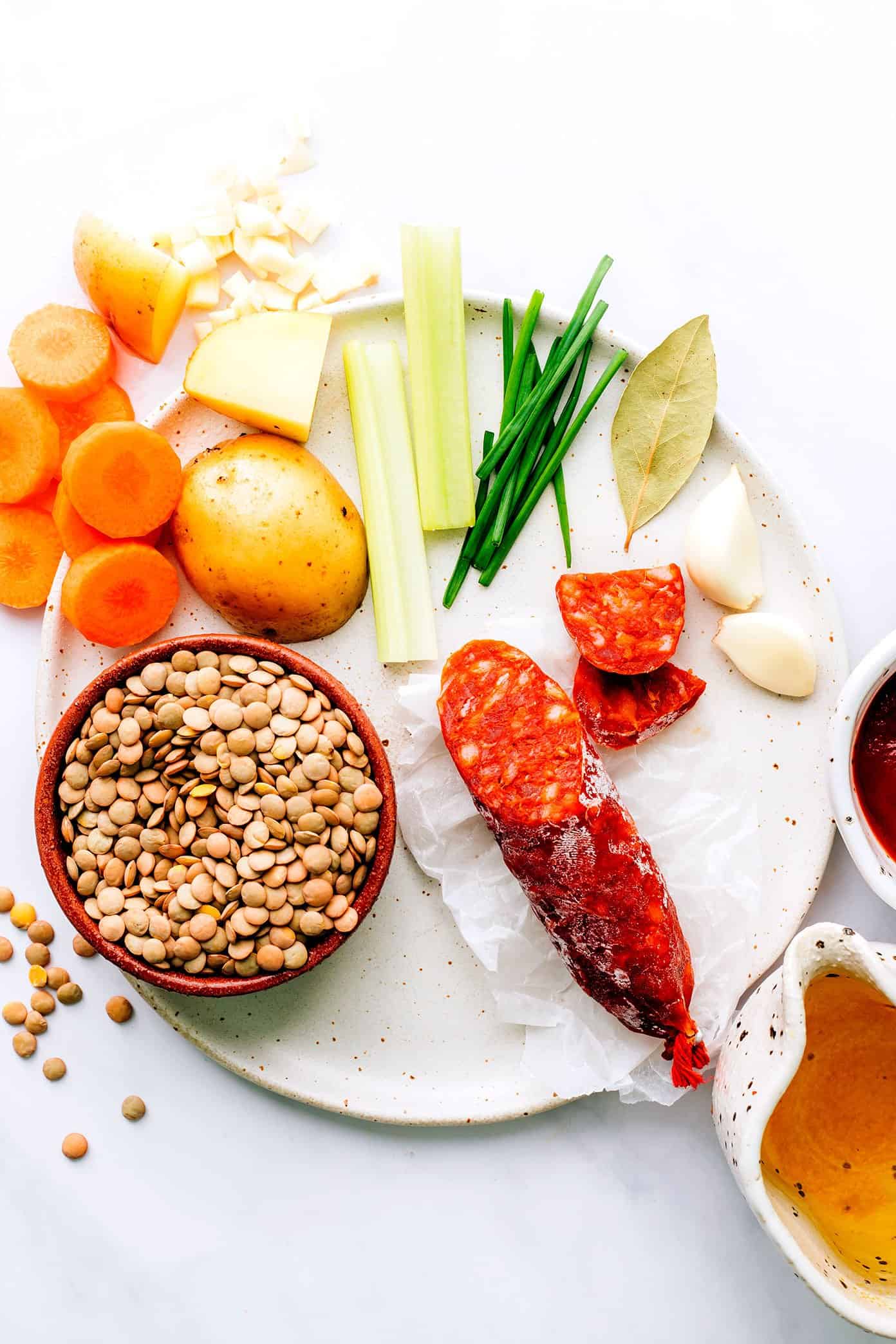 Spanish Chorizo Lentil Soup ingredients on plate, including carrots, potato, garlic, celery, chives, bay leaf, garlic, tomato paste and broth